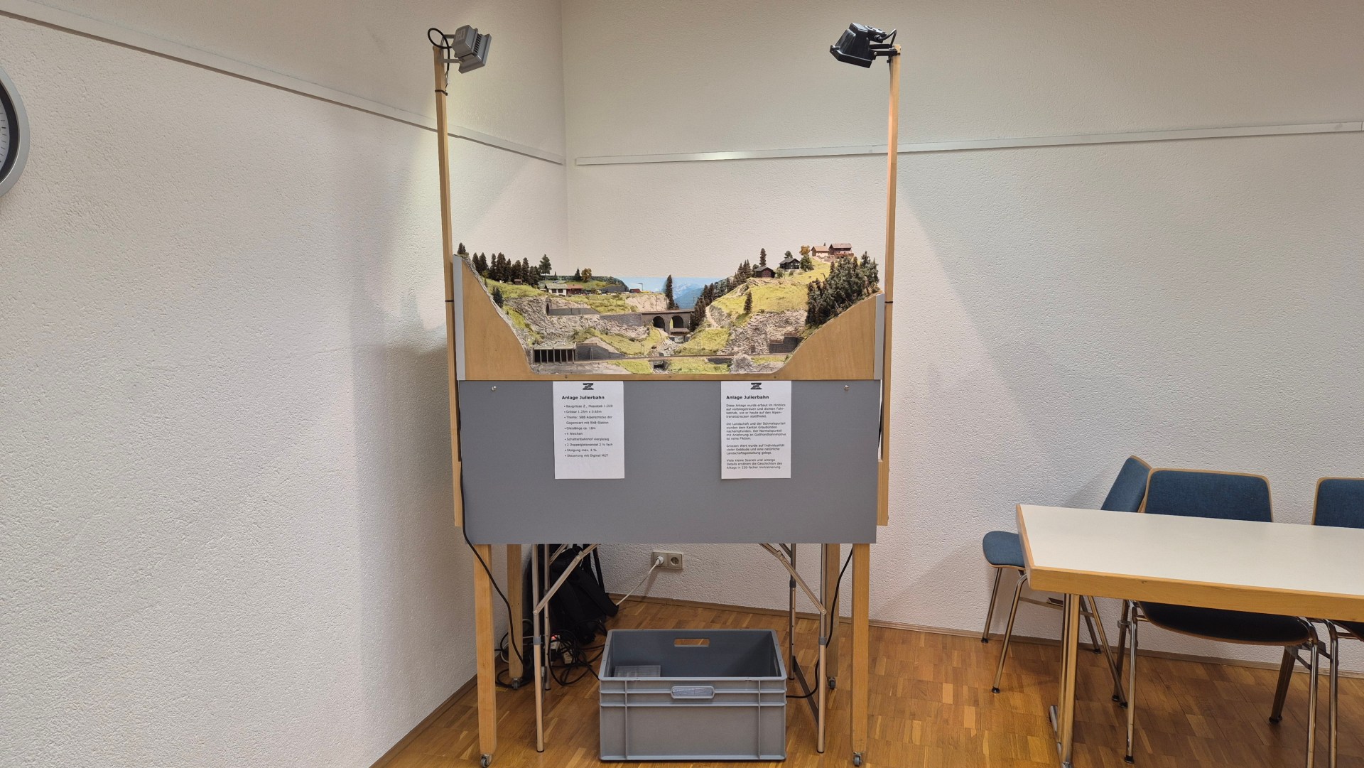 Modellbahnanlage in einer Ausstellung mit beschildertem Hintergrund, umgeben von Tischen und Stühlen.
