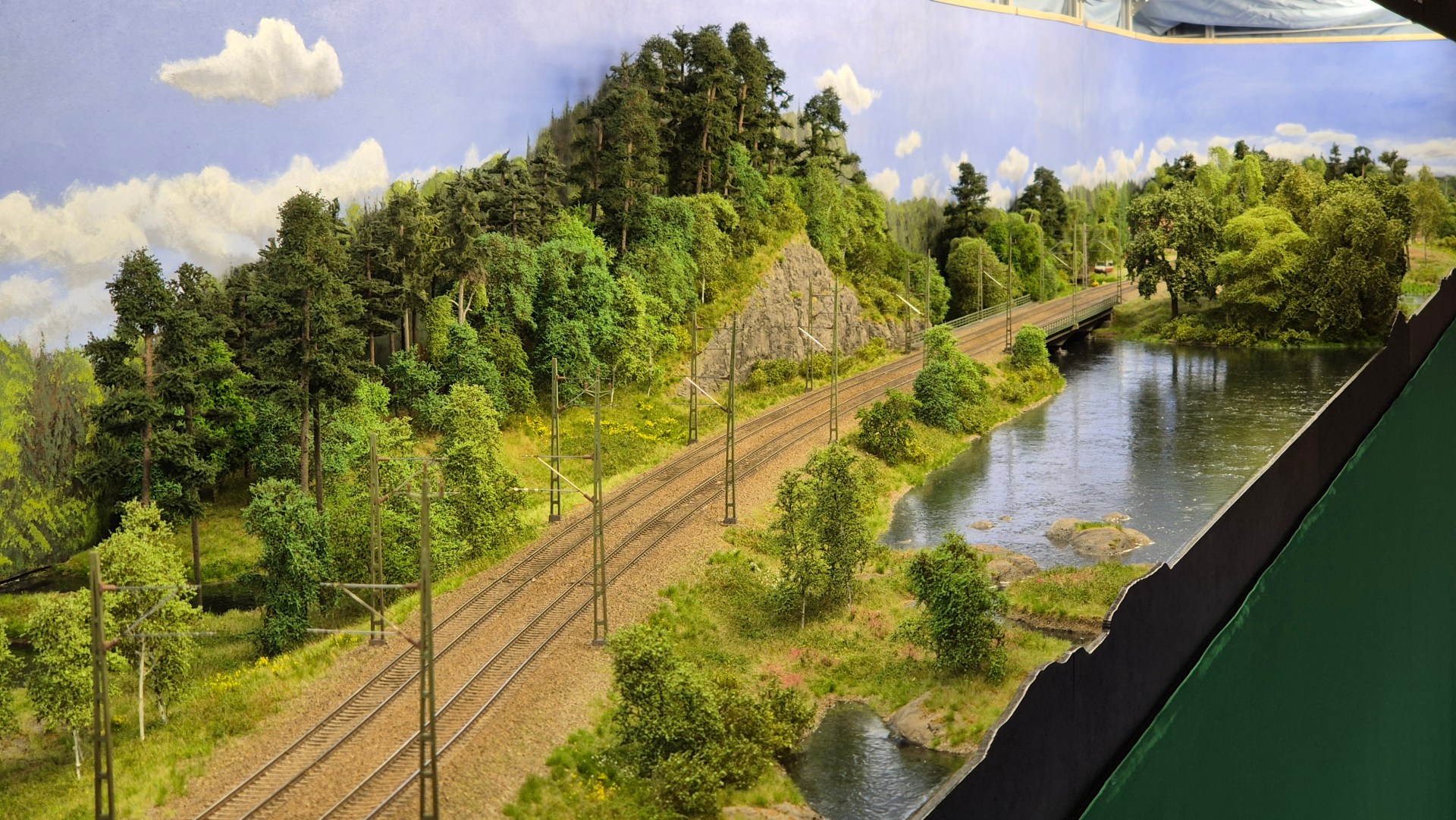 A detailed model railway landscape featuring lush greenery, trees, and a river alongside the tracks. The scene includes electric poles and a bridge in the background, showcasing a miniature representation of a realistic railway environment.