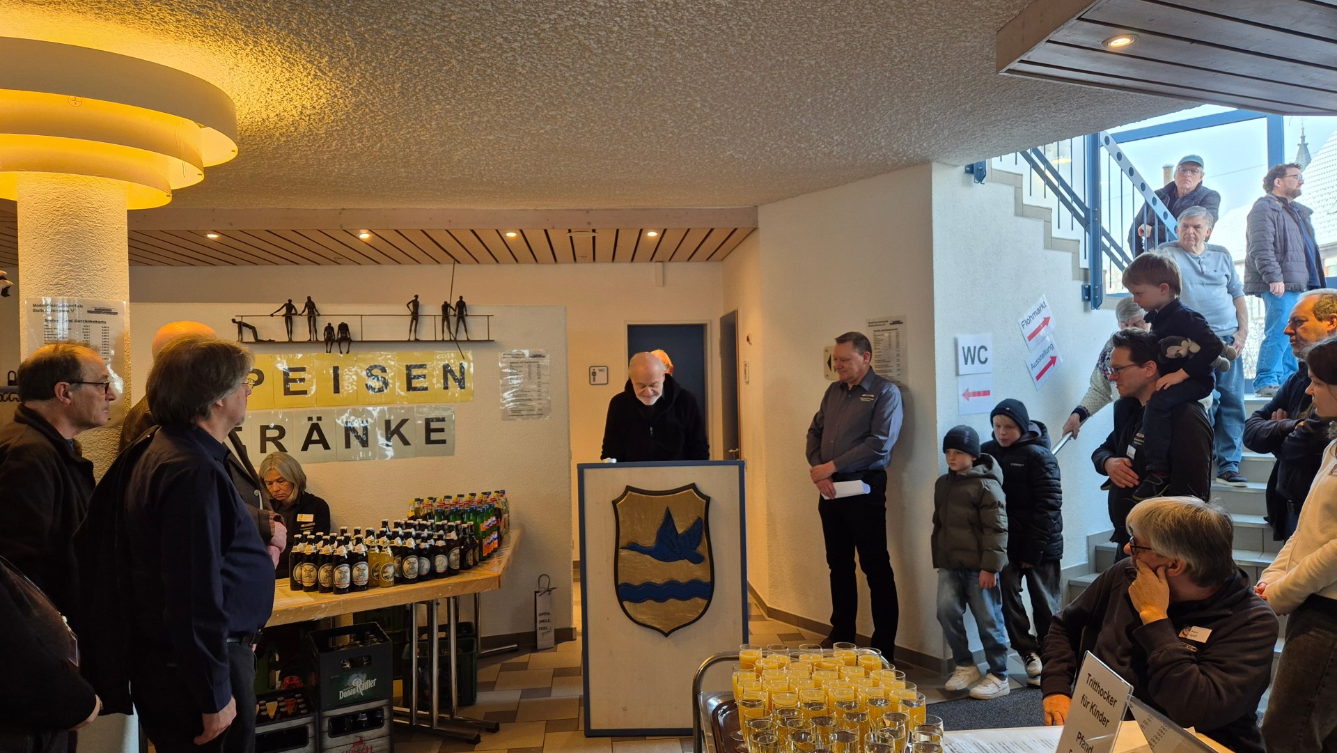 Ein Veranstaltungsraum mit Menschen, die aufmerksam einem Redner zuhören. Im Vordergrund steht ein Tisch mit Getränken und Snacks, während im Hintergrund eine große Gruppe von Besuchern an einer Treppe steht. Ein Schild mit der Aufschrift "WC" ist sichtbar.