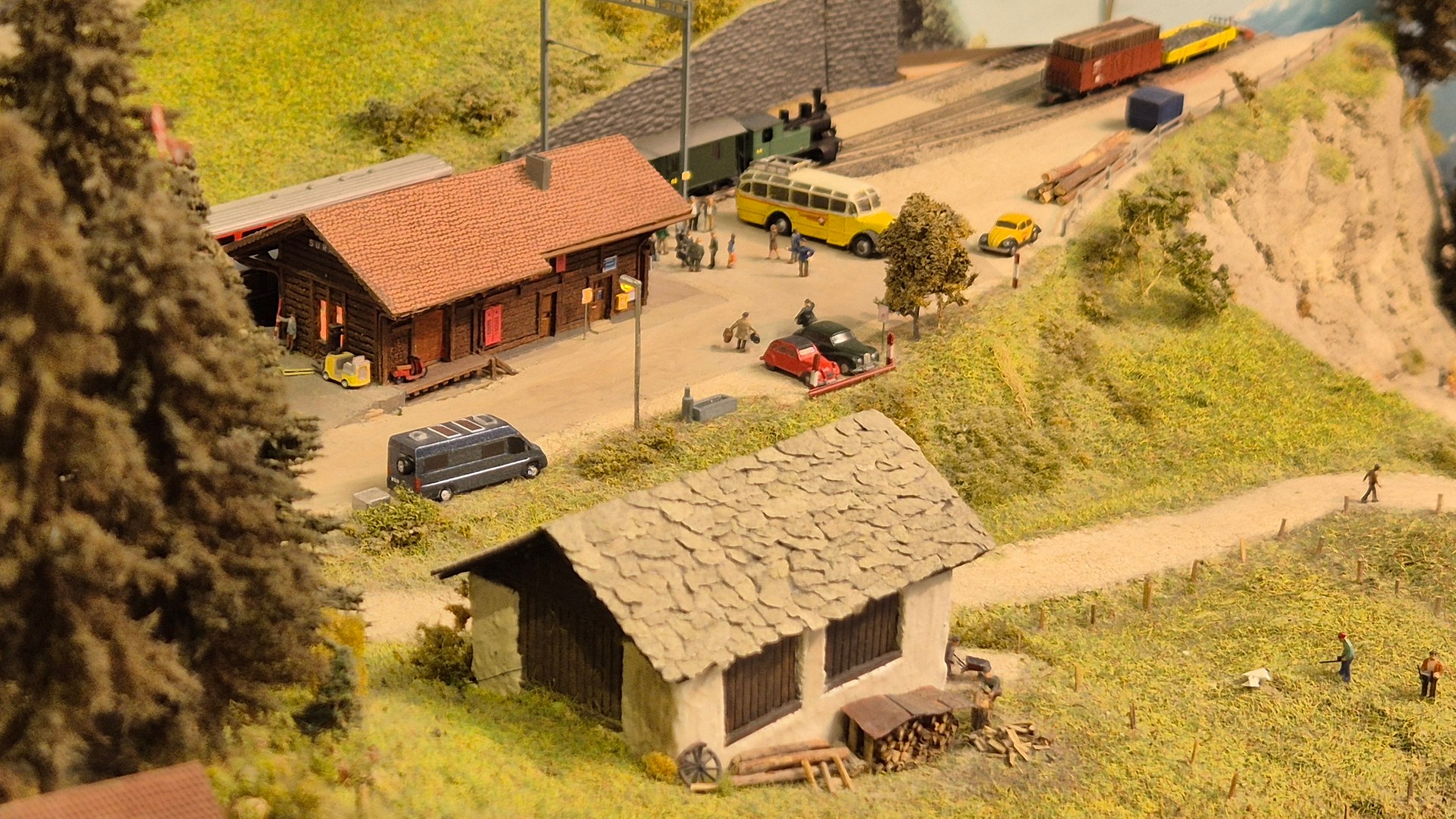 Ansicht einer Modellbahnanlage mit einem Bahnhofsgebäude, mehreren Fahrzeugen, und Figuren in einer ländlichen Szenerie.
